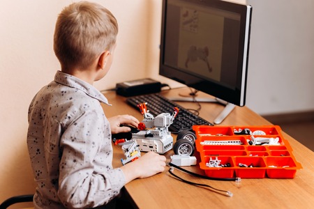 Smart boy dressed in gray shirt makes a robot from the robotic constructor at the desk with computer...