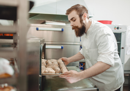 Young charismatic baker puts a baking tray with breads rolls in the oven in the bakeryの写真素材