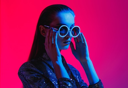 Fashion girl with long hair and round sunglasses in a black shining dress poses in neon light in the studioの写真素材
