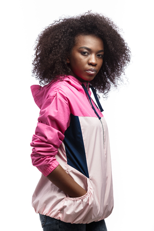 Saucy young curly brown-haired girl dressed in the pink sport jacket stands at the white background in the studioの写真素材