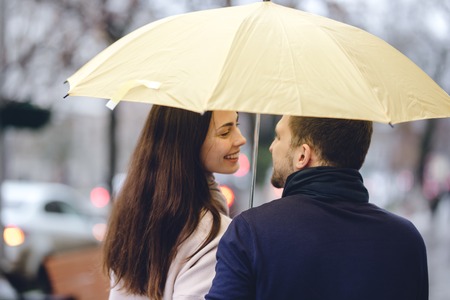 Beautiful couple, guy and his girlfriend dressed in casual clothes stand under the umbrella and look at each other on the street in the rainの写真素材