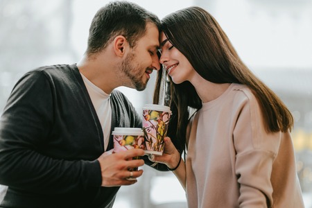 Loving couple is siting face to face on the background of window and holding cups in their hands.の写真素材