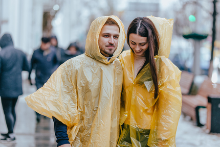 Happy guy and his girlfriend dressed in yellow raincoats are walking on the street in the rainの写真素材