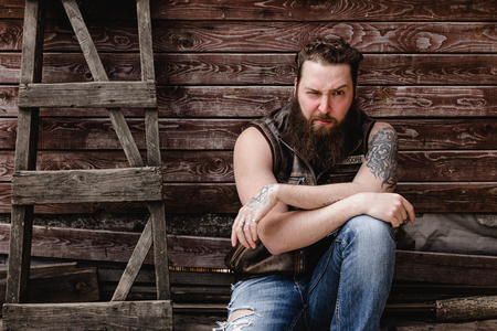 Strong brutal man with a beard and tattoos on his hands dressed in leather vest and jeans  sits on a wooden wall background next to a wooden ladder outsideの写真素材