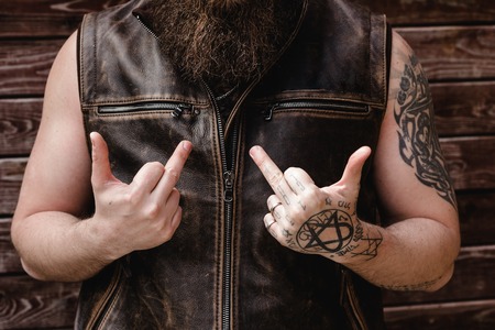 Strong brutal man with a beard and tattoos on his hands dressed in leather vest shows middle fingersの写真素材