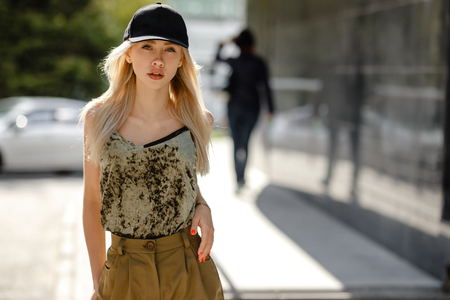 Young blonde girl dressed in fashionable trousers and top khaki color and black cap on the head poses in the streetの写真素材