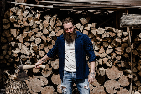 Brutal man with a beard dressed in casual clothes with tattoos on his hands holds the ax sitting near the stacked woodの写真素材