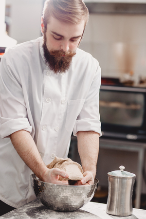 Professional baker kneads dough in an iron bowl on the table in the kitchen of the bakeryの写真素材