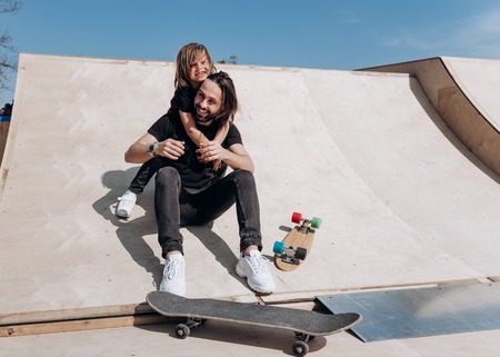 Happy young father and his son dressed in the stylish casual clothes are sitting in an embrace together on the slide next to the skateboards in a skate park at the sunny warm dayの写真素材