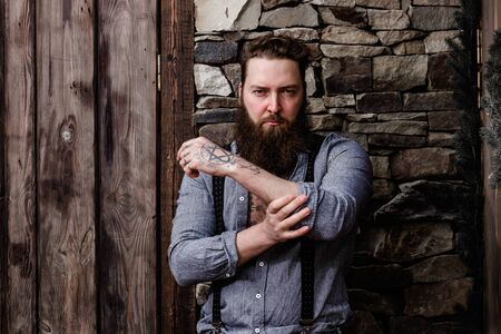 Brutal strong man with a beard and tattoos on his hands dressed in stylish casual clothes stands on the background of stone wall and wooden doorの写真素材