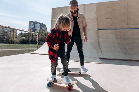 Father and his little son dressed in the casual clothes ride skateboards in a skate park with slides outside at the sunny dayの写真素材