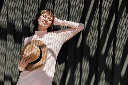 Stylish girl in a gently pink dress and straw hat standing with the straw hat  in her hand next to a dark metal fence in the sunlight with reflected shadowsの写真素材