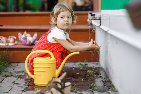 A little girl dressed in a sundress holds her hands under running tap water sitting next to the watering can in the garden next to the house on the summer dayの写真素材