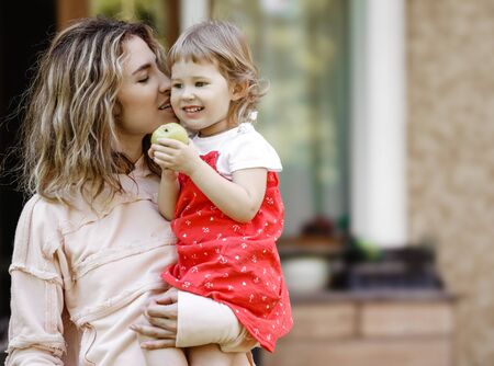 Young mother holds her little daughter on her hands and kisses her sweet cheek in the garden on a warm summer dayの写真素材