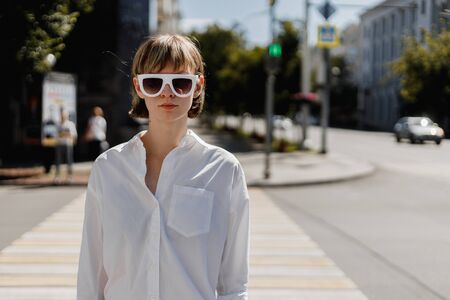 Stylish young woman in white sunglasses dressed in white shirt is standing in the city street on a summer sunny dayの写真素材