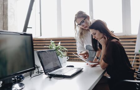 Two young beautiful women dressed according to office dreskod are working together with laptop at the desk in a light modern officeの写真素材