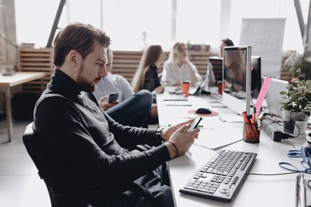 The guy dressed in casual office style clothes is working sitting at desk with computer next to the colleagues in the modern officeの写真素材