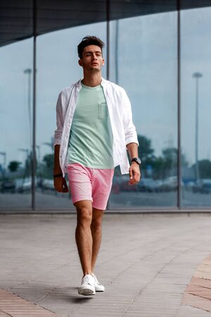Stylish guy dressed in trendy summer clothes in casual style posing against the background of a glass building on a summer dayの写真素材