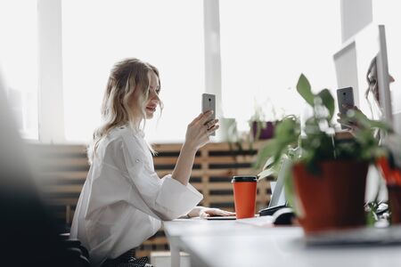 Young beautiful woman dressed in white shirt is working with phone sitting at the desk with a laptop...