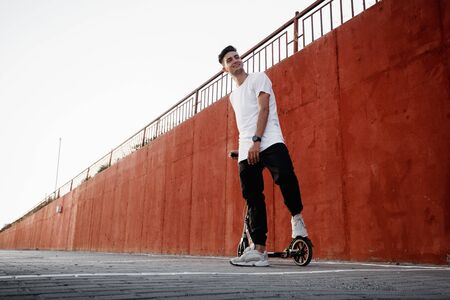 Young guy dressed in jeans and t-shirt stands with a scooter against a painted concrete wall on the...