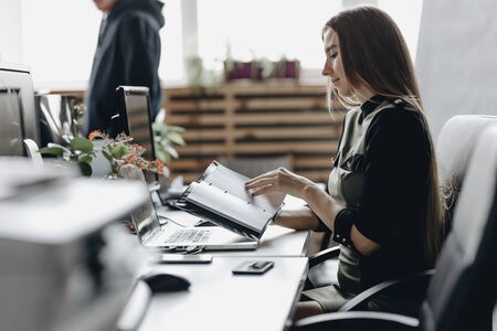 Young brunette girl dressed in office style clothes is sitting at the desk and keeps the notebook in...