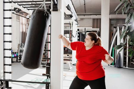 Fat girl in the sportswear is boxing punching bag in the gym