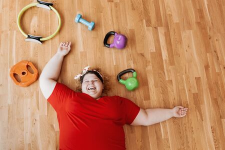 Laughing fat girl dressed in the sportswear and with a bandage on her head is laying on the floor next to the sports equipmentの写真素材