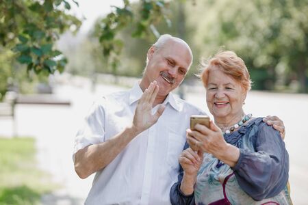Old man and old woman are using together one phone and smiling while walking in a park on a warm dayの写真素材