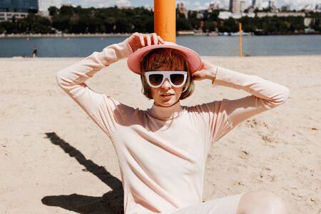 Stylish girl in white sunglasses, a gently pink dress and pink visor sits on a sandy beach leaning...