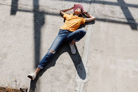 The girl in yellow t-shirt, jeans and pink visor is laying on a concrete slab on a sunny day outside