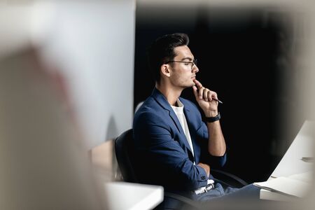 Architect in glasses dressed in blue checkered jacket sits in the armchair near the desk with laptop in the officeの写真素材