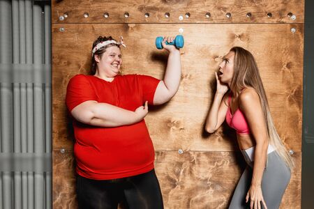Fat and funny girl dressed in the sportswear with dumbbells in hands stands next to the surprised...