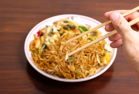 Hand holding chopsticks to eat fried noodle with vegetables on wooden tableの写真素材