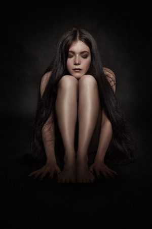 pensive young naked woman sitting in darkの写真素材