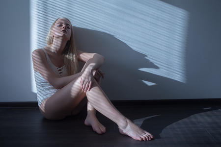 sad girl in linen sits on floor in moonlight with blinds toned imageの写真素材