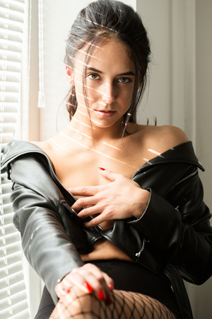 sad pensive girl in leather jacket near window with blindsの写真素材