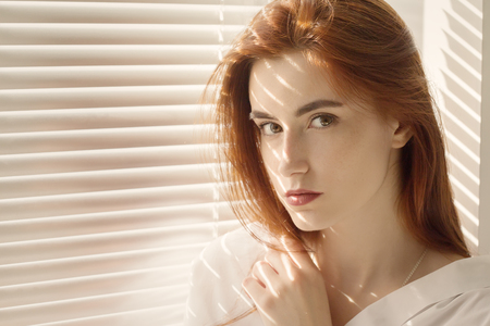 sad beautiful woman sitting at window with blinds in sunlight, looking at cameraの写真素材