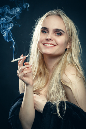 happy young blond woman with bare shoulders on dark blue background smoking smilingの写真素材
