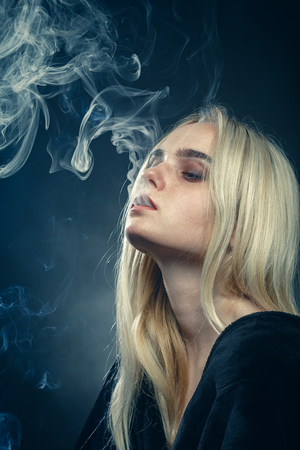 serious young blond woman on dark blue background smokingの写真素材