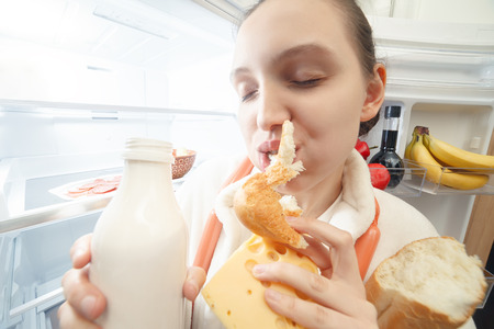 hungry fun woman eating food near opened refrigeratorの写真素材