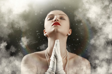 sensual luxury young woman with closed eyes in white gloves on black sky background with stars and clouds prays under light raysの写真素材