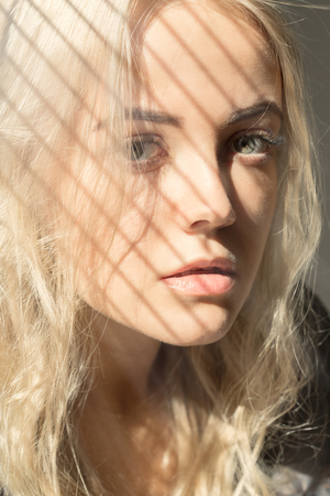sad blonde woman in sun light with blinds shadow looking at cameraの写真素材