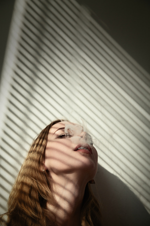 sad young woman in sunlight with blinds shadows smoking cigaretteの写真素材