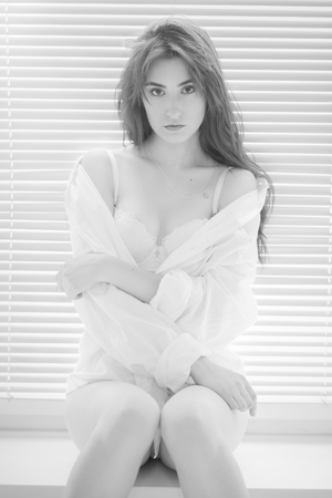 sad pensive girl in white blouse sitting on window in sunlight with blinds, monochromeの写真素材