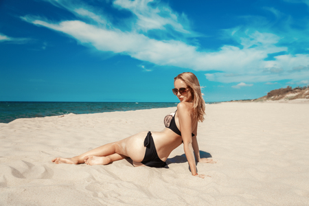 single happy young blond woman in swimsuit and sunglasses at sand sea beach tanning, sitting, toned imageの写真素材