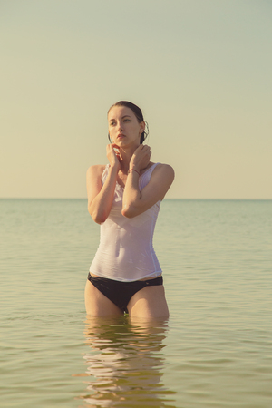 beautiful young woman in transparent wet shirt stand in calm tropical sea under golden sunset light toned imageの写真素材