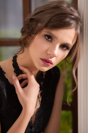 beautiful pensive girl in black dress near window looking at cameraの写真素材