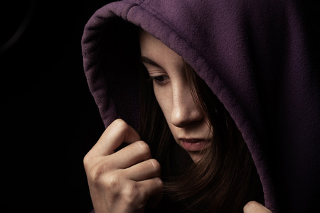 scared girl in hood looking down on black backgroundの写真素材
