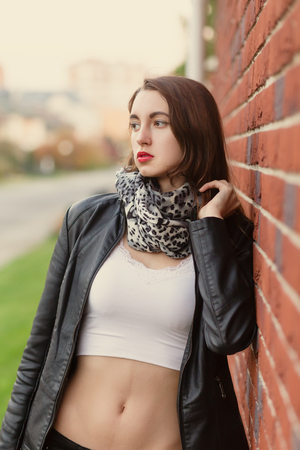 attractive sad young woman in leather coat standing near bricks wallの写真素材