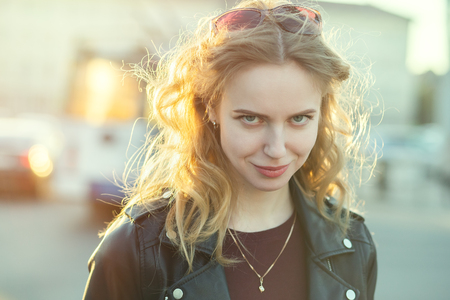 fun shy happy blond girl in leather coat looking at camera on city street smiling under sun rays toned imageの写真素材